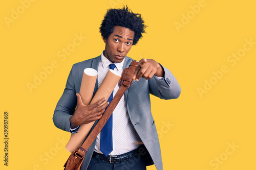 Handsome african american man with afro hair holding paper blueprints pointing with finger to the camera and to you, confident gesture looking serious