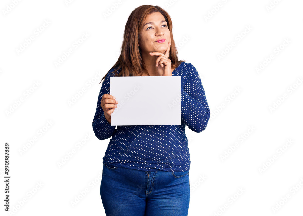 Middle age latin woman holding paper banner with blank space serious face thinking about question with hand on chin, thoughtful about confusing idea Middle age latin woman holding paper banner with blank space serious face thinking about question with hand on chin, thoughtful about confusing idea