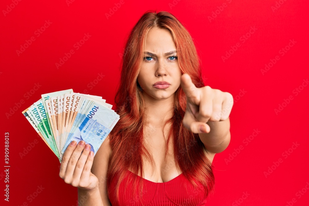 Young beautiful redhead woman holding south korean won banknotes pointing with finger to the camera and to you, confident gesture looking serious Young beautiful redhead woman holding south korean won banknotes pointing with finger to the camera and to you, confident gesture looking serious