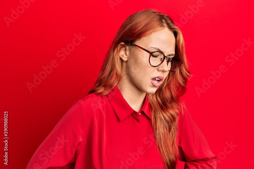 Young beautiful redhead woman wearing casual clothes and glasses over red background with hand on stomach because nausea, painful disease feeling unwell. ache concept.