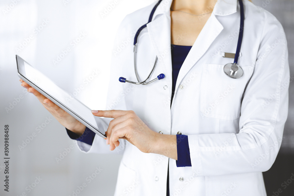 Unknown woman-doctor is holding a tablet computer in her hands, while standing in a clinic. Female physician with a stethoscope in her office, close-up. Perfect medical service in a hospital. Medicine Unknown woman-doctor is holding a tablet computer in her hands, while standing in a clinic. Female physician with a stethoscope in her office, close-up. Perfect medical service in a hospital. Medicine