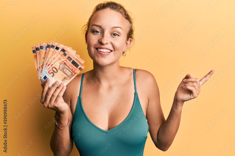 Beautiful caucasian woman holding bunch of 50 euro banknotes smiling happy pointing with hand and finger to the side Beautiful caucasian woman holding bunch of 50 euro banknotes smiling happy pointing with hand and finger to the side