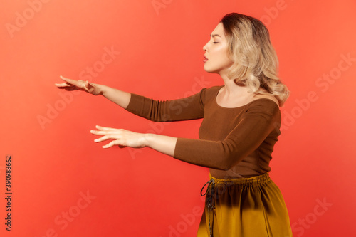 Side view portrait of blonde beautiful woman in brown blouse standing with closed eyes and outstretched hands, trying to move forward blind. Indoor studio shot isolated on red background