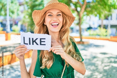 Young beautiful blonde woman on vacation wearing summer hat smiling happy. Standing with smile on face holding paper with like message at street of city.