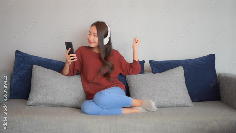 Young happy asian woman enjoys in music from smartphone with wireless headphones on couch in living room, full frame slow motion Young happy asian woman enjoys in music from smartphone with wireless headphones on couch in living room, full frame slow motion