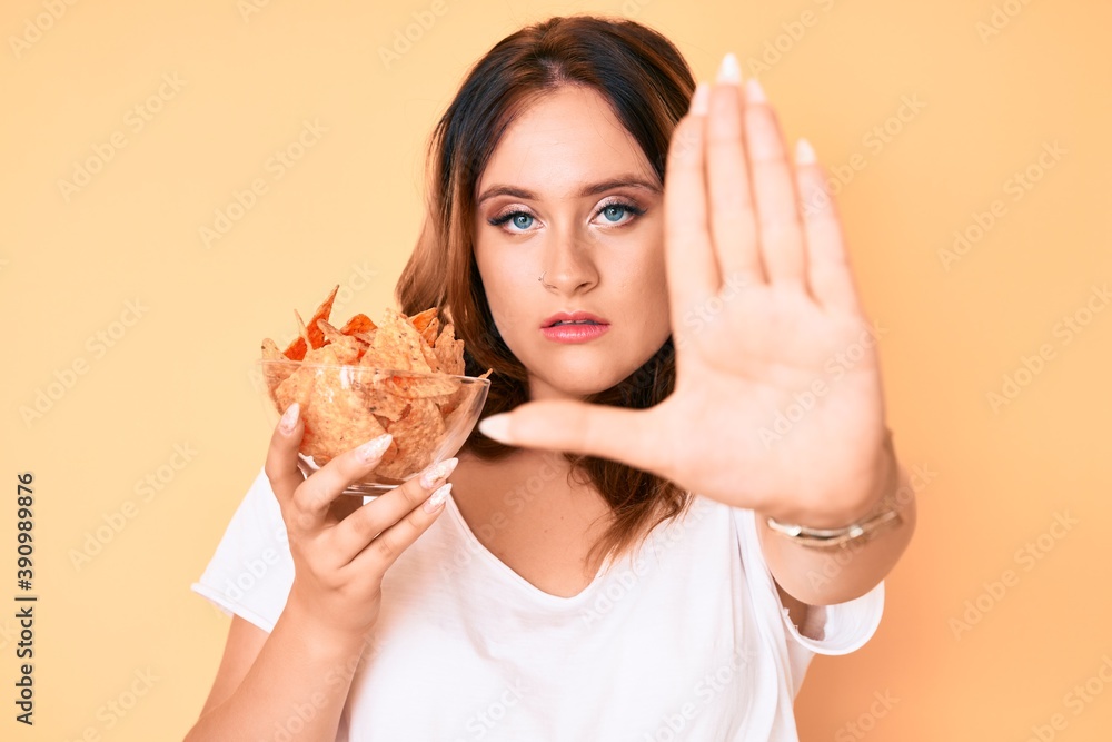 Young beautiful caucasian woman holding nachos potato chips with open hand doing stop sign with serious and confident expression, defense gesture Young beautiful caucasian woman holding nachos potato chips with open hand doing stop sign with serious and confident expression, defense gesture