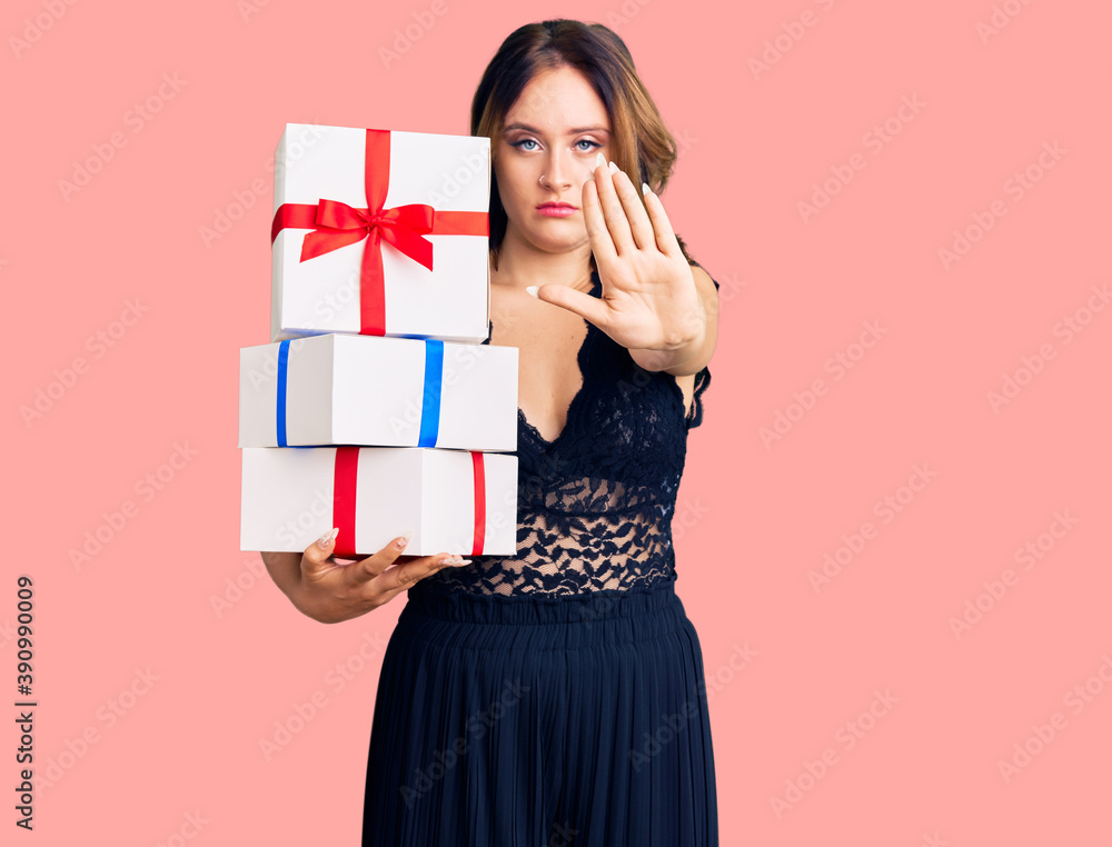 Young beautiful caucasian woman holding gifts with open hand doing stop sign with serious and confident expression, defense gesture Young beautiful caucasian woman holding gifts with open hand doing stop sign with serious and confident expression, defense gesture