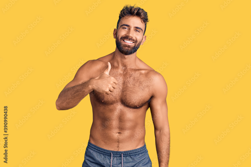 Young hispanic man wearing swimwear shirtless doing happy thumbs up gesture with hand. approving expression looking at the camera showing success.