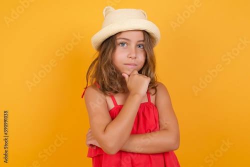 Thoughtful smiling young Caucasian girl standing against yellow background keeps hand under chin, looks directly at camera, listens something with interest. Youth concept.