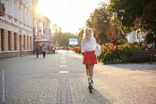 Young blond woman, riding away from camera on electric scooter in city center during sunset. Summer leisure activity. Traveler exploring the town, spending time outdoors.