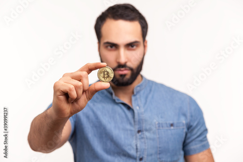 Serious bearded man holding in fingers and showing golden coin bitcoin crypto currency, electronic virtual money. Indoor studio shot isolated on white background