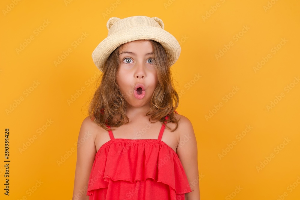 Dreamy young Caucasian girl wearing hat and red dress standing against yellow wall, with shocked expression. Dreamy young Caucasian girl wearing hat and red dress standing against yellow wall, with shocked expression.
