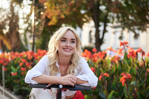 Young blond woman, standing with electric scooter in city center near flowerbeds. Three-quarter portrait of female, wearing red and white outfit. Summer leisure activity. Spending free time outdoors.