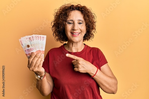 Beautiful middle age mature woman holding 10 colombian pesos banknotes smiling happy pointing with hand and finger
