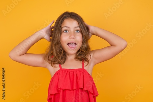 young Caucasian girl standing against yellow background keeps hands on chest feeling shocked and scared, mouth widely opened, stares at camera saying: Who, me?