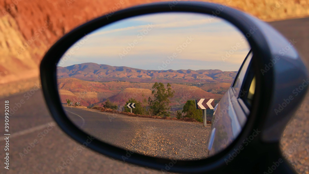 Beautifully red colored foothills of the stunning Atlas Mountains in the evening sun with paved street and road signs near Ouzoud, Morocco viewed in the side mirror of a small car. Focus on center.