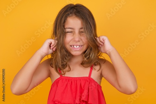 Stop making this annoying sound! Unhappy stressed out Caucasian young girl standing against yellow background making worry face, plugging ears with fingers, irritated with loud noise.