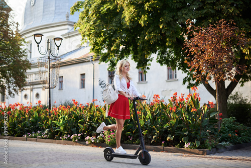 Young blond woman, riding on electric scooter in city center near flowerbeds. Full-length action portrait of female. Summer leisure activity. Spending free time outdoors. Traveler exploring the town.
