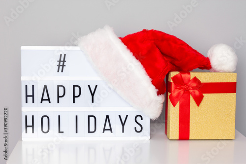 Lightbox with happy holidays hashtag text, Santa hat and gift box on white table