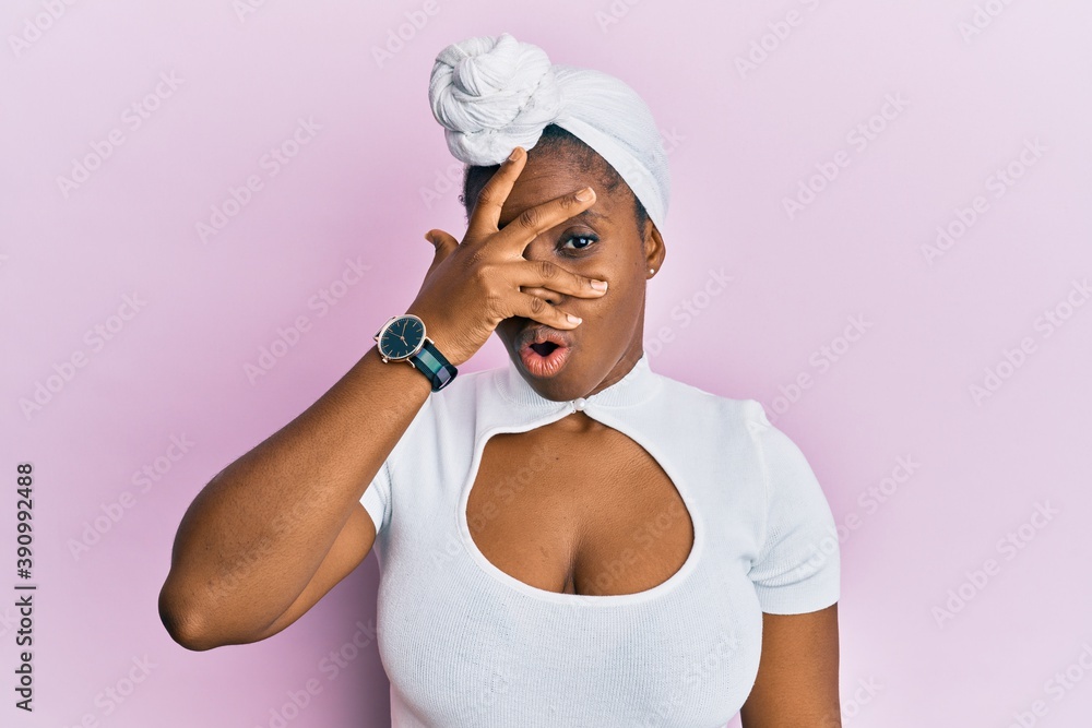 Young african woman wearing hair turban over pink background peeking in shock covering face and eyes with hand, looking through fingers afraid Young african woman wearing hair turban over pink background peeking in shock covering face and eyes with hand, looking through fingers afraid