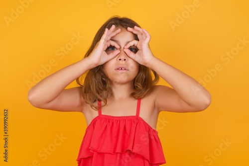 Playful excited Caucasian young girl standing against yellow background showing Ok sign with both hands on eyes, pretending to wear spectacles.