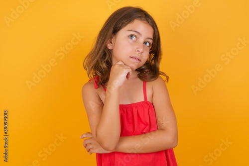 Face expressions and emotions. Thoughtful young Caucasian girl standing against yellow background holding hand under his head, having doubtful look.