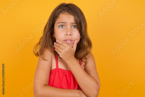 young Caucasian girl standing against yellow background covering mouth with hands scared from something or someone bitting nails