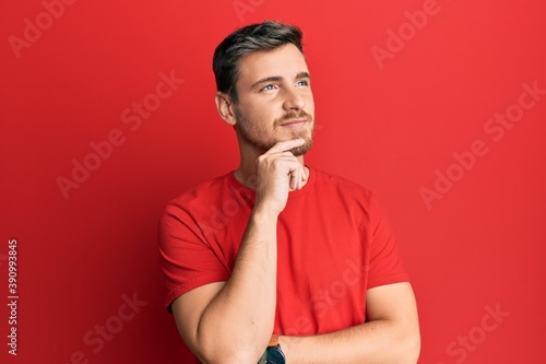 Handsome caucasian man wearing casual red tshirt with hand on chin thinking about question, pensive expression. smiling with thoughtful face. doubt concept.