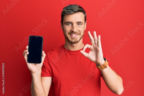 Handsome caucasian man holding smartphone showing screen doing ok sign with fingers, smiling friendly gesturing excellent symbol