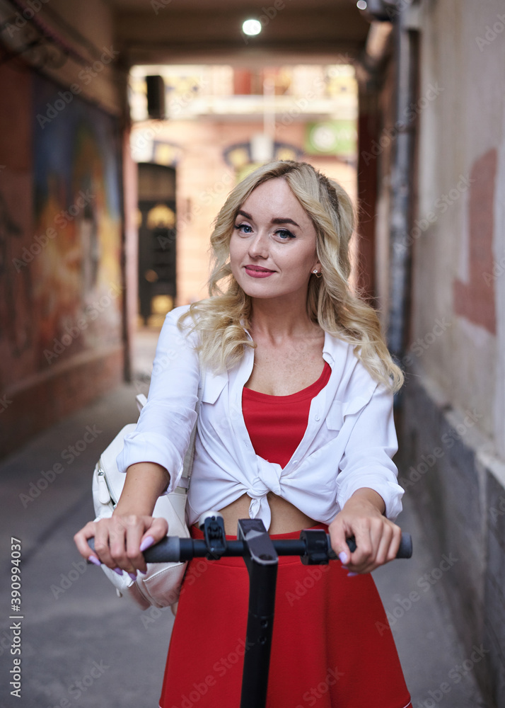 Young blond woman, standing with electric scooter in city center near old building wall. Female, wearing red and white outfit, posing.Summer leisure activity.Traveler exploring the town.