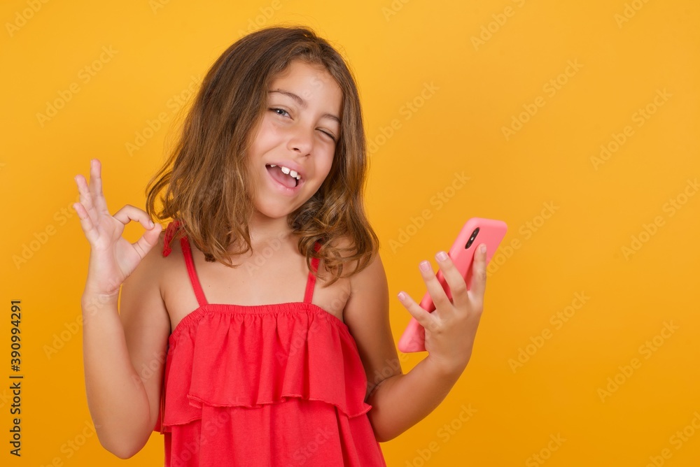 Happy Caucasian young girl standing against yellow background sending a message on his smartphone or taking a selfie and making ok sign with his hand. Happy Caucasian young girl standing against yellow background sending a message on his smartphone or taking a selfie and making ok sign with his hand.