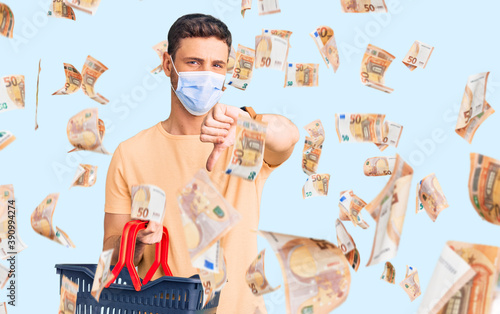Handsome young man with bear wearing shopping basket and medical mask with angry face, negative sign showing dislike with thumbs down, rejection concept