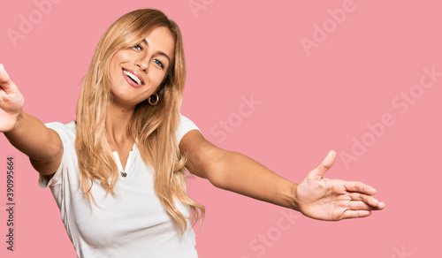 Beautiful blonde young woman wearing casual white tshirt looking at the camera smiling with open arms for hug. cheerful expression embracing happiness.