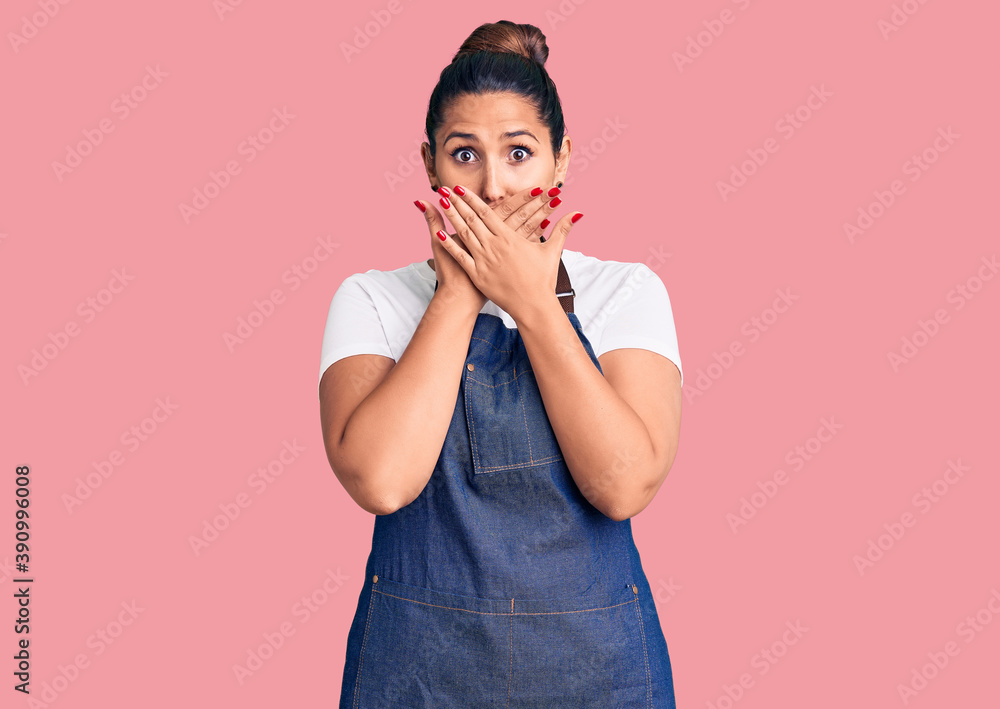 Young beautiful brunette woman wearing apron shocked covering mouth with hands for mistake. secret concept.