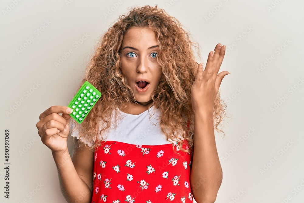 Beautiful caucasian teenager girl holding birth control pills scared and amazed with open mouth for surprise, disbelief face Beautiful caucasian teenager girl holding birth control pills scared and amazed with open mouth for surprise, disbelief face