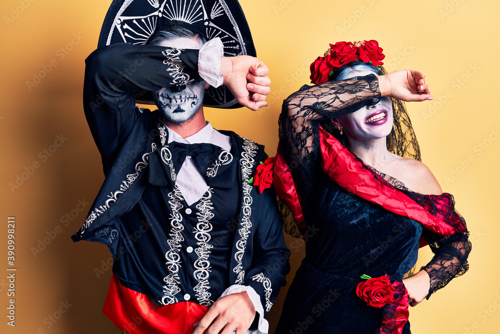 Young couple wearing mexican day of the dead costume over yellow covering eyes with arm smiling cheerful and funny. blind concept. Young couple wearing mexican day of the dead costume over yellow covering eyes with arm smiling cheerful and funny. blind concept.
