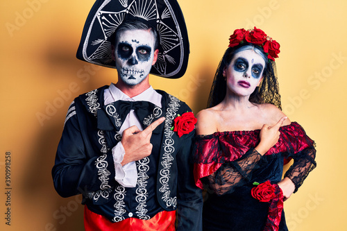 Young couple wearing mexican day of the dead costume over yellow pointing with hand finger to the side showing advertisement, serious and calm face