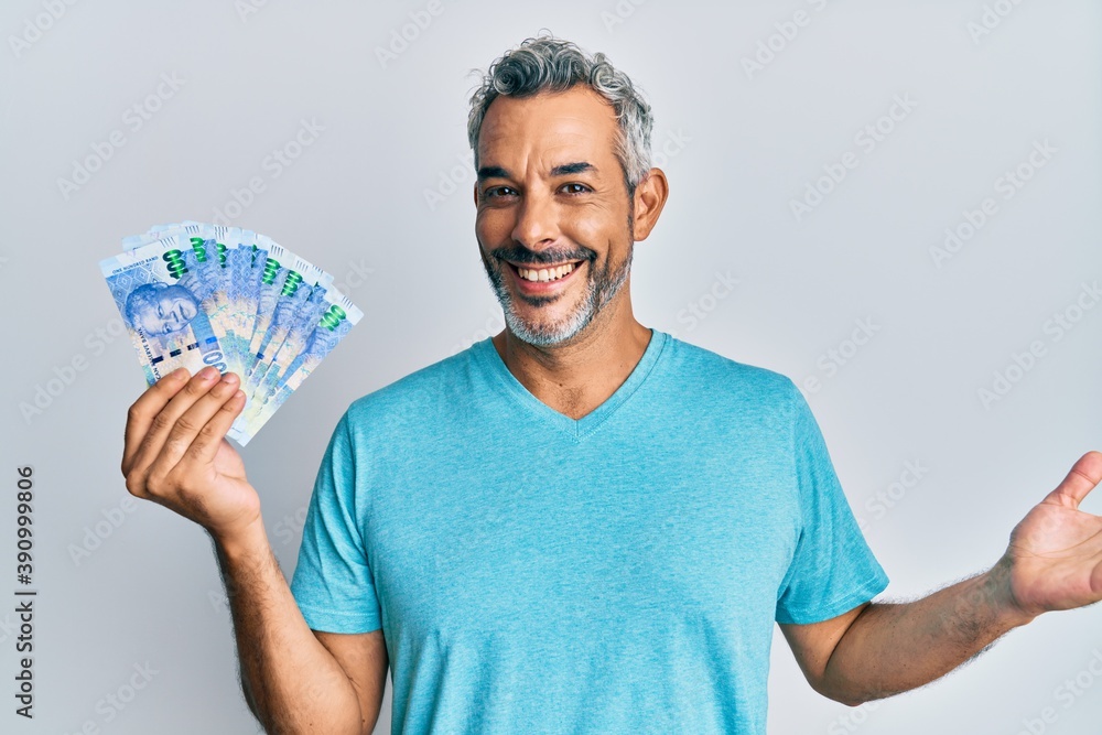 Middle age grey-haired man holding south african rands banknotes celebrating achievement with happy smile and winner expression with raised hand Middle age grey-haired man holding south african rands banknotes celebrating achievement with happy smile and winner expression with raised hand