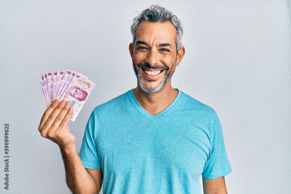 Middle age grey-haired man holding mexican pesos looking positive and happy standing and smiling with a confident smile showing teeth Middle age grey-haired man holding mexican pesos looking positive and happy standing and smiling with a confident smile showing teeth