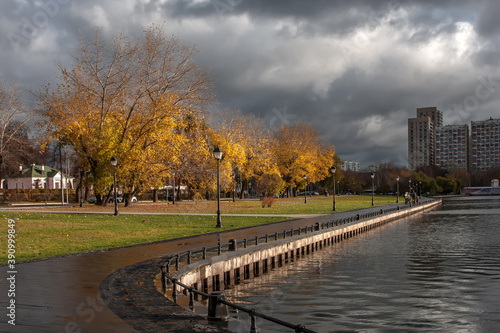 Beautiful and empty embankment of the autumn pond. Yellow trees, clouds in the sky. City autumn landscape. Wet asphalt.
