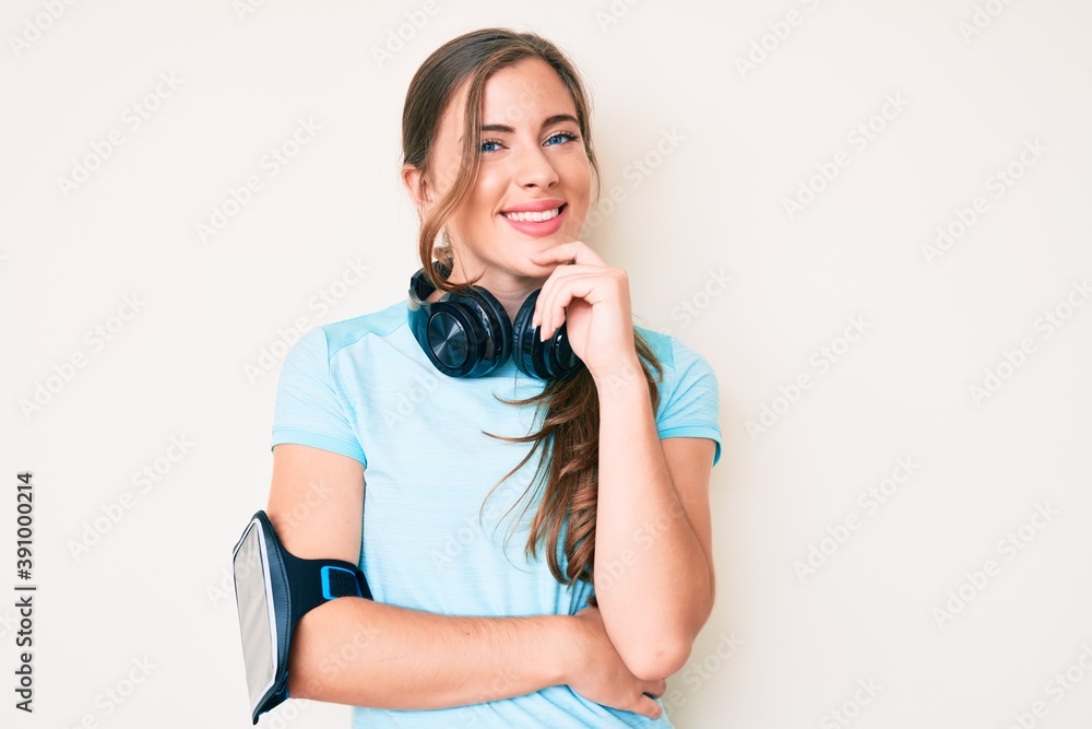 Beautiful young caucasian woman wearing gym clothes and using headphones looking confident at the camera with smile with crossed arms and hand raised on chin. thinking positive. Beautiful young caucasian woman wearing gym clothes and using headphones looking confident at the camera with smile with crossed arms and hand raised on chin. thinking positive.