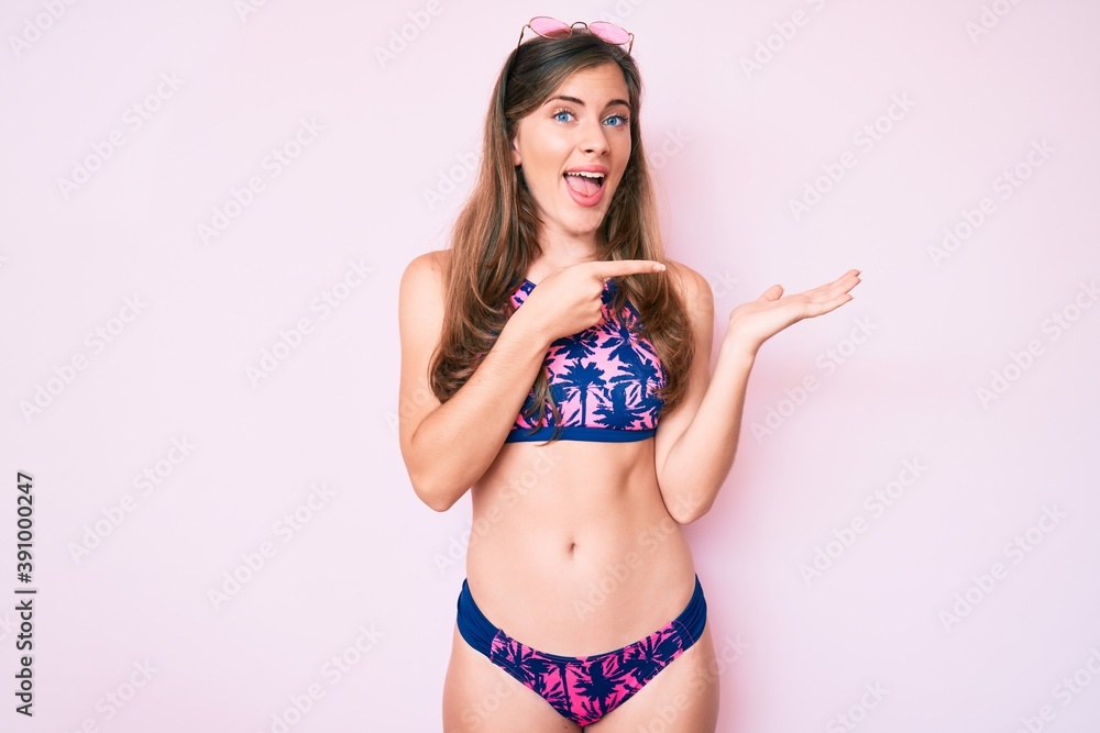 Naklejka premium Beautiful young caucasian woman wearing bikini and sunglasses amazed and smiling to the camera while presenting with hand and pointing with finger.