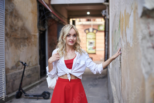 Young blond woman, leaning on old building wall in city center. Three-quarter portrait of female, wearing red and white outfit, posing. Summer leisure activity. Traveler exploring the town. © Natalia