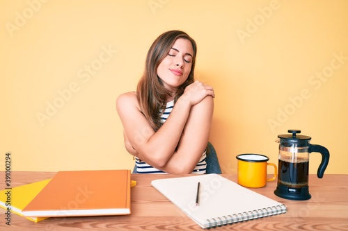 Beautiful young caucasian woman sitting on the table stuying for university hugging oneself happy and positive, smiling confident. self love and self care photo