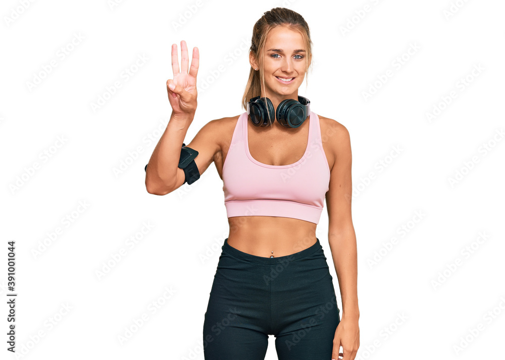 Young blonde woman wearing gym clothes and using headphones showing and pointing up with fingers number three while smiling confident and happy. Young blonde woman wearing gym clothes and using headphones showing and pointing up with fingers number three while smiling confident and happy.