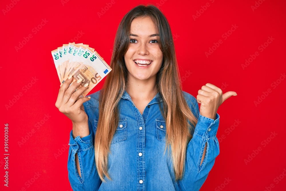 Beautiful caucasian woman holding 50 euro banknotes pointing thumb up to the side smiling happy with open mouth Beautiful caucasian woman holding 50 euro banknotes pointing thumb up to the side smiling happy with open mouth