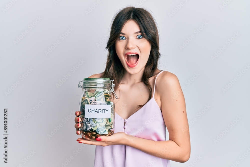 Young beautiful caucasian girl holding charity jar with money celebrating crazy and amazed for success with open eyes screaming excited. Young beautiful caucasian girl holding charity jar with money celebrating crazy and amazed for success with open eyes screaming excited.