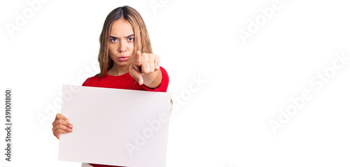 Young beautiful blonde woman holding blank empty banner pointing with finger to the camera and to you, confident gesture looking serious photo