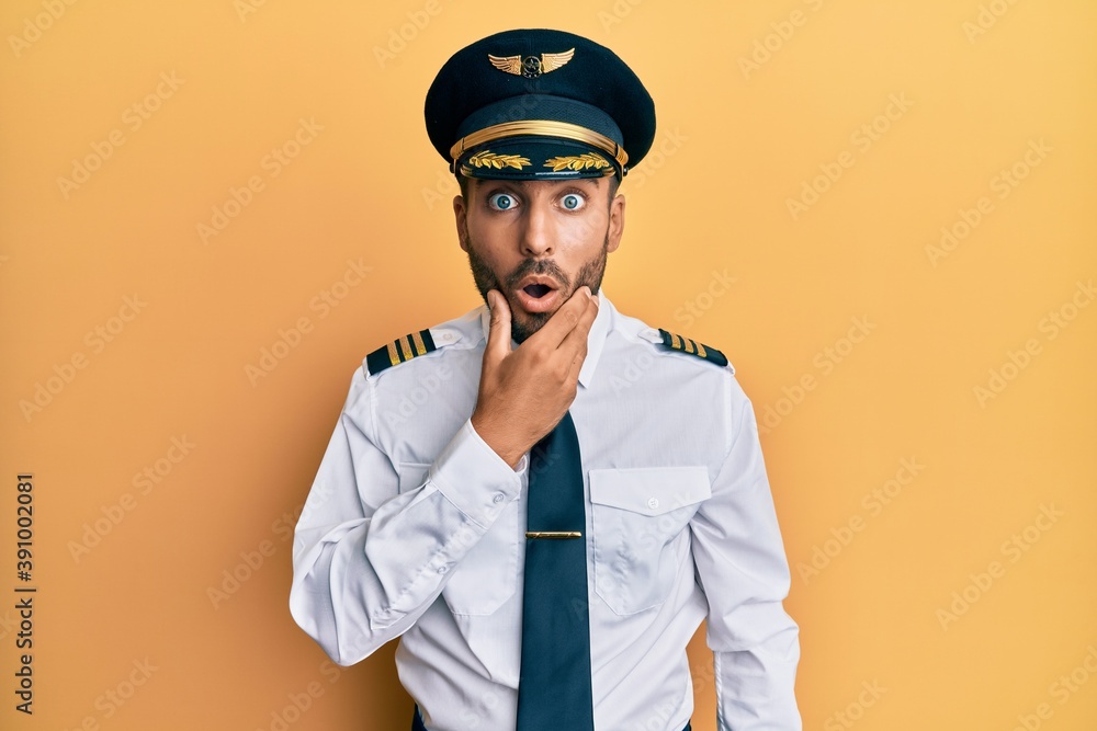 Handsome hispanic man wearing airplane pilot uniform looking fascinated with disbelief, surprise and amazed expression with hands on chin Handsome hispanic man wearing airplane pilot uniform looking fascinated with disbelief, surprise and amazed expression with hands on chin