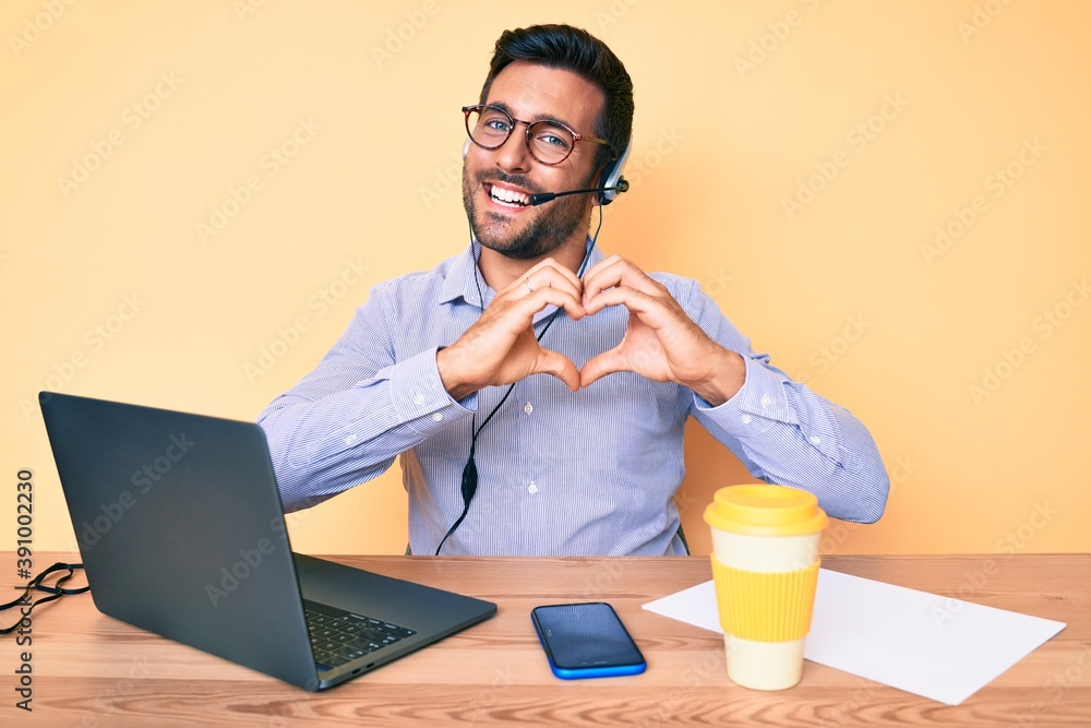 Young hispanic man sitting at the desk wearing operator headset at the call center office smiling in love showing heart symbol and shape with hands. romantic concept. Young hispanic man sitting at the desk wearing operator headset at the call center office smiling in love showing heart symbol and shape with hands. romantic concept.